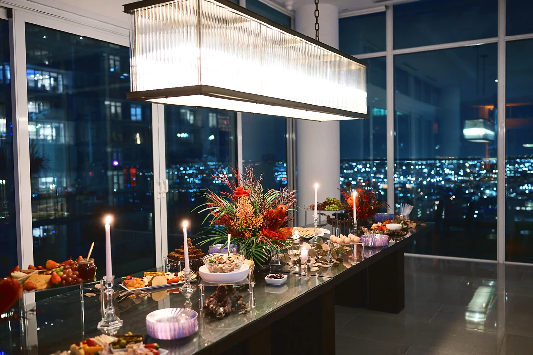A candlelit grazing table under a glass chandelier with floor-to-ceiling windows and the city at night