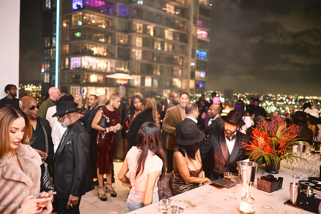 Guests in hats and evening wear at the rooftop bar beside an arrangement of red and orange flowers